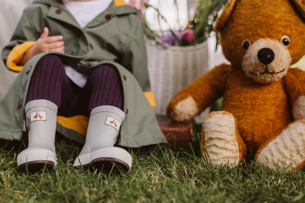 Grey waterproof wellies worn by a child sitting next to a teddy bear. 