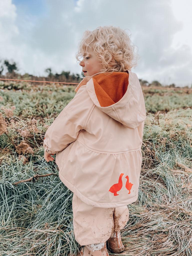 Baby girl with blonde curly hair on a farm wearing a pink waterproof rain jacket with an adjustable hood. 