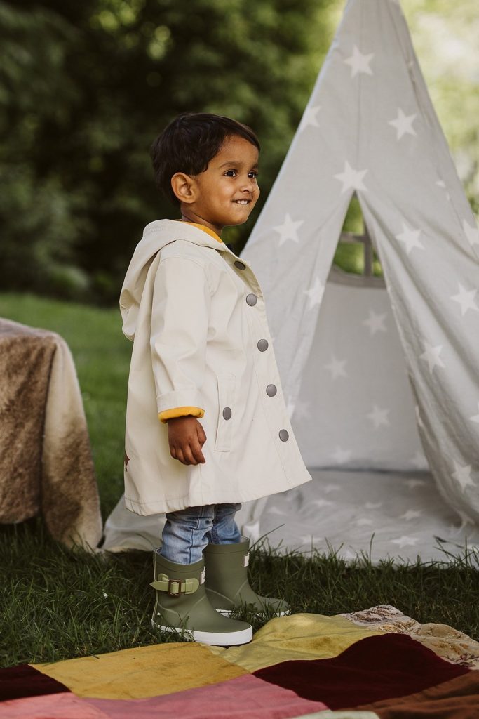 Young boy smiling, wearing green waterproof wellies and a cream rain jacket outside. 