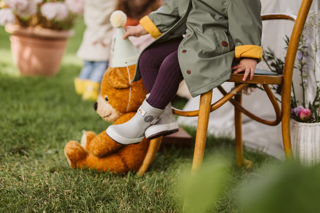 A child sitting on a wooden chair outside wearing a green waterproof jacket and grey wellies next to a teddy