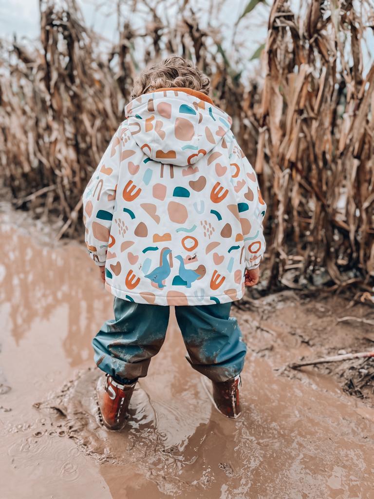 A toddler boy with curly hair is wearing a geo-print pink and white rain jacket and muddy wellies on a mucky and wet surface. 